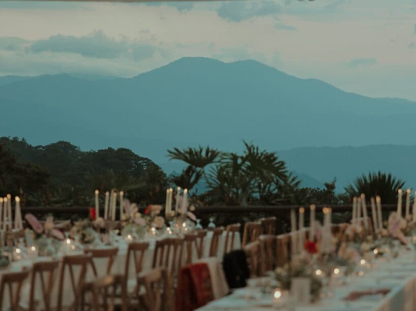 A long banquet table set with tapered candles and pale florals on an open-air terrace, looking out over layered blue mountain ridges at dusk
