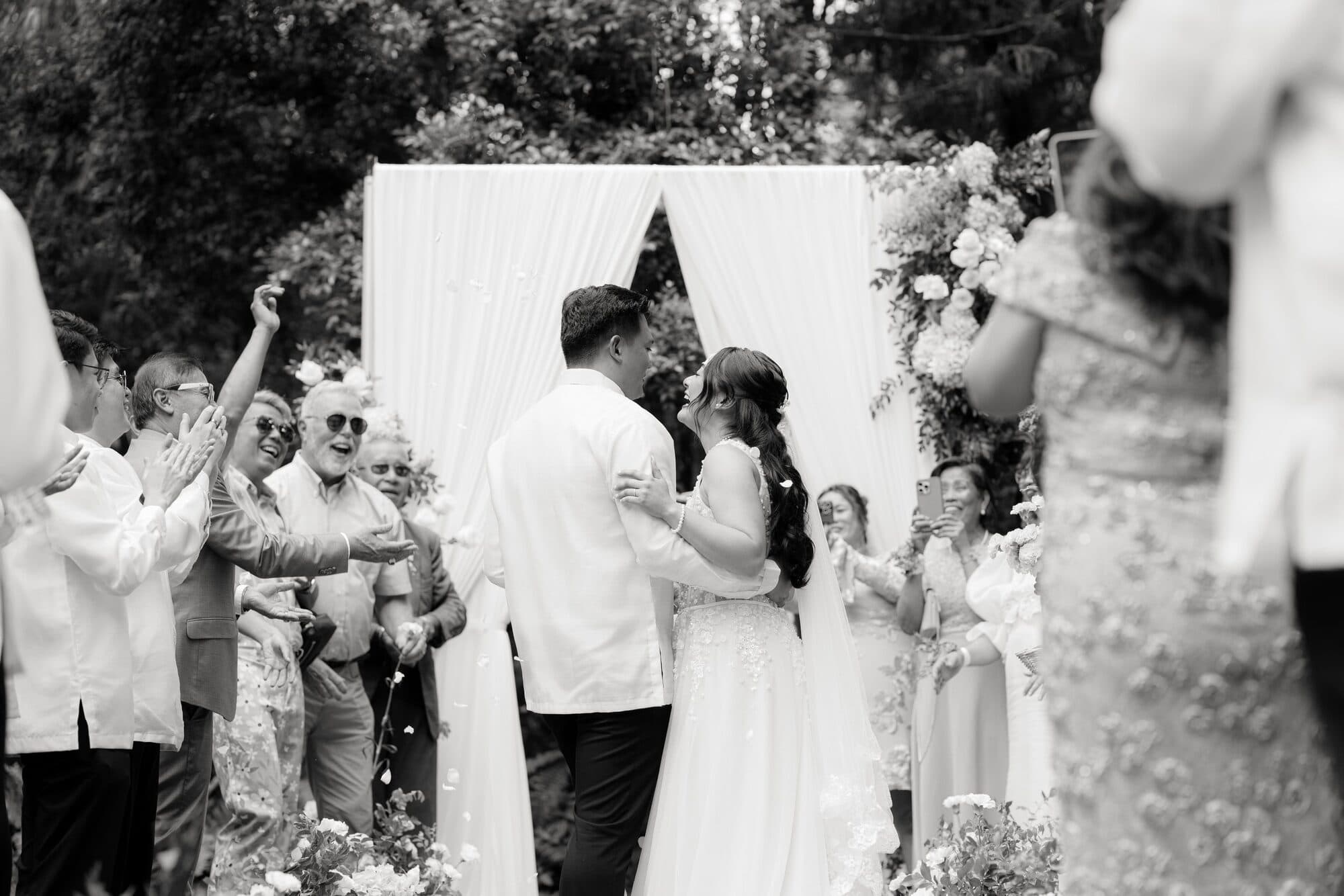 The couple embracing at the ceremony arch while guests throw petals around them