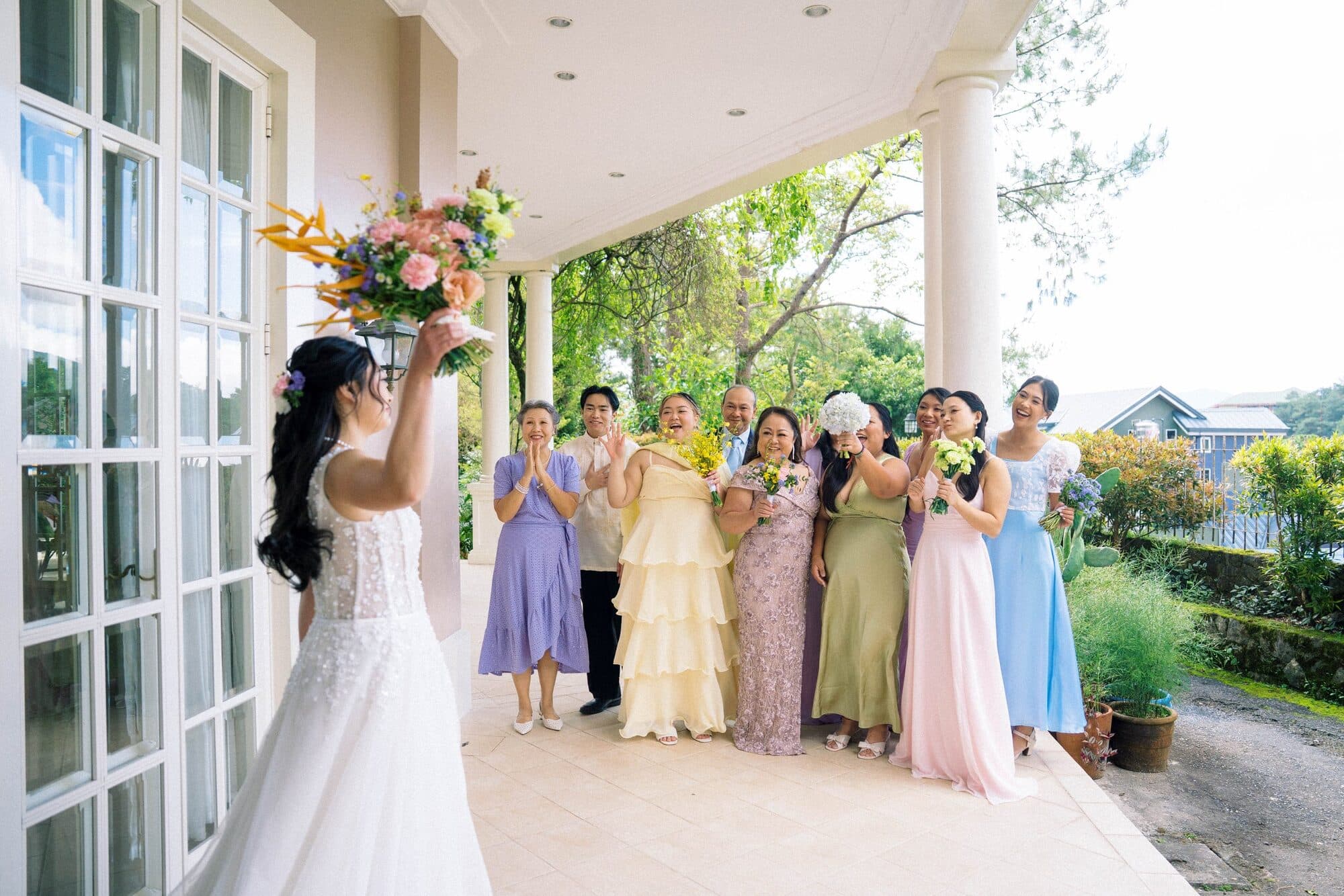 The bride raising her bouquet on the veranda toward her mothers and bridesmaids gathered in pastel gowns
