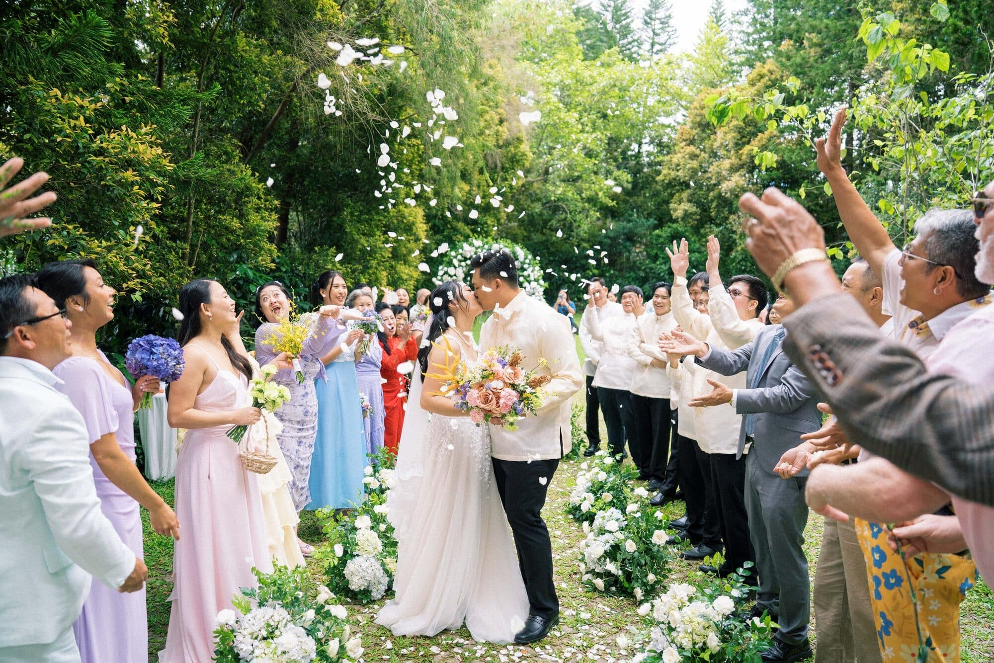 The couple kissing beneath a shower of petals as their guests cheer along the garden aisle