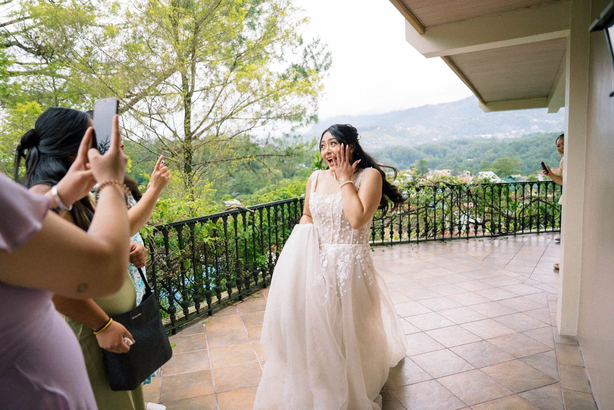 The bride on the balcony above the Baguio hills, hand to her cheek in surprise as friends photograph her