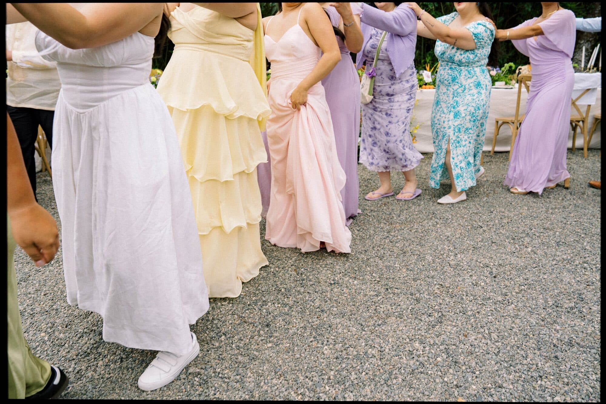 Guests in pastel dresses dancing together in a circle on the gravel