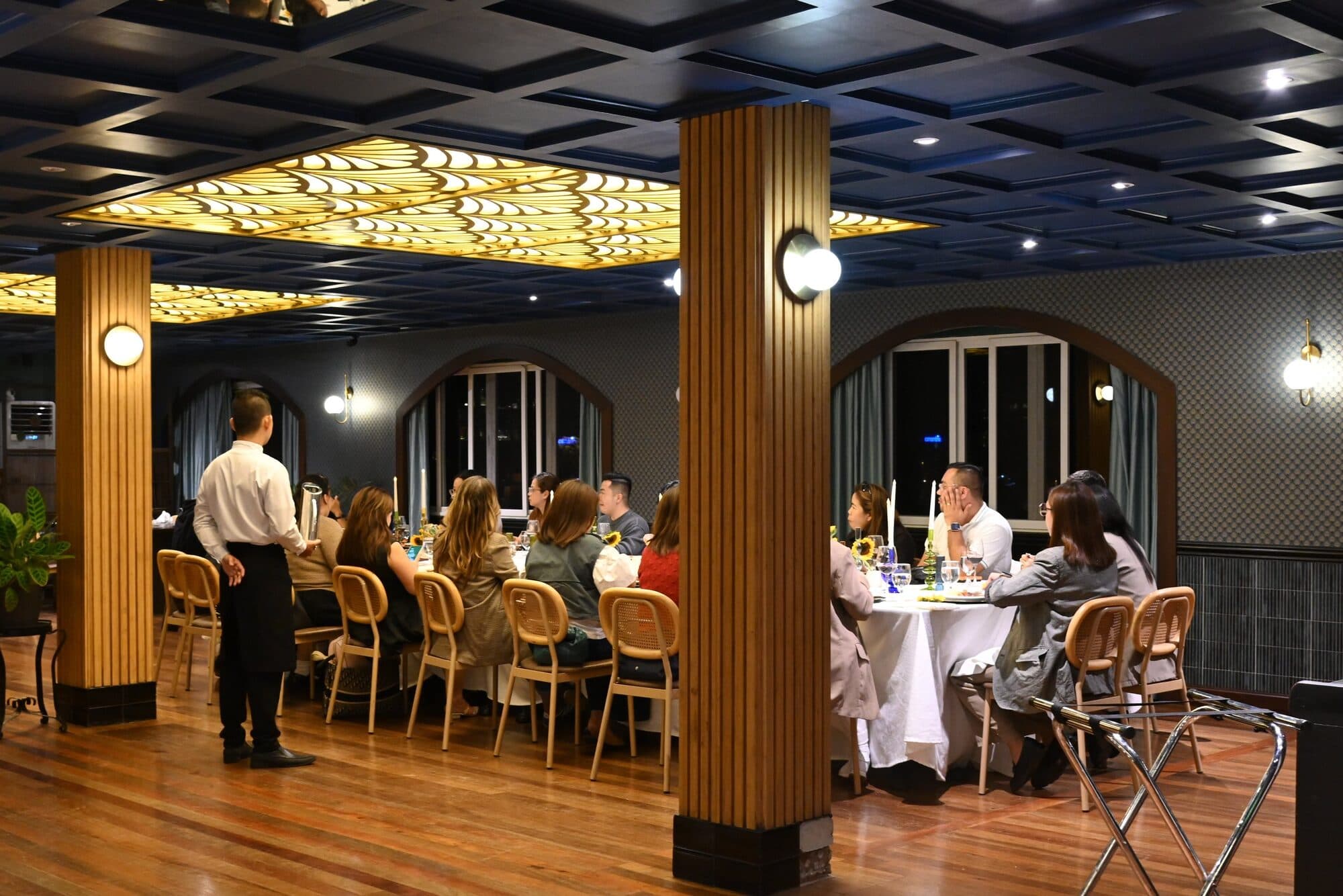 Guests seated along a long white-clothed table in Mario’s dining room, under its carved gold ceiling panel and wooden columns