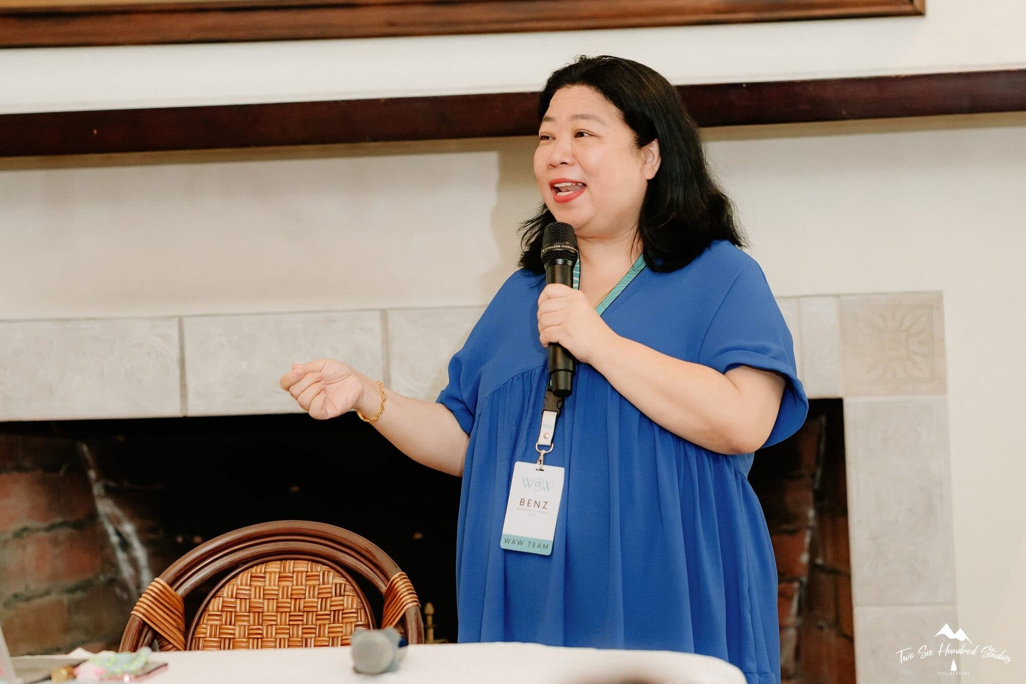 Benz Co-Rana speaking into a microphone in front of the fireplace, gesturing as she talks