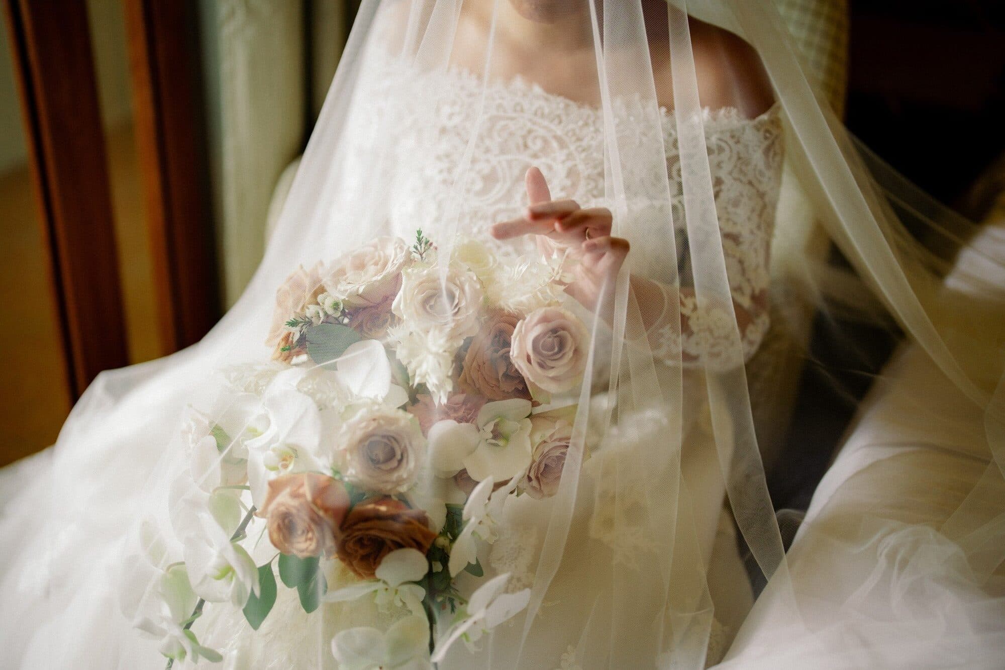 A bride under a sheer veil holding a bouquet of blush roses, orchids and eucalyptus against her lace gown
