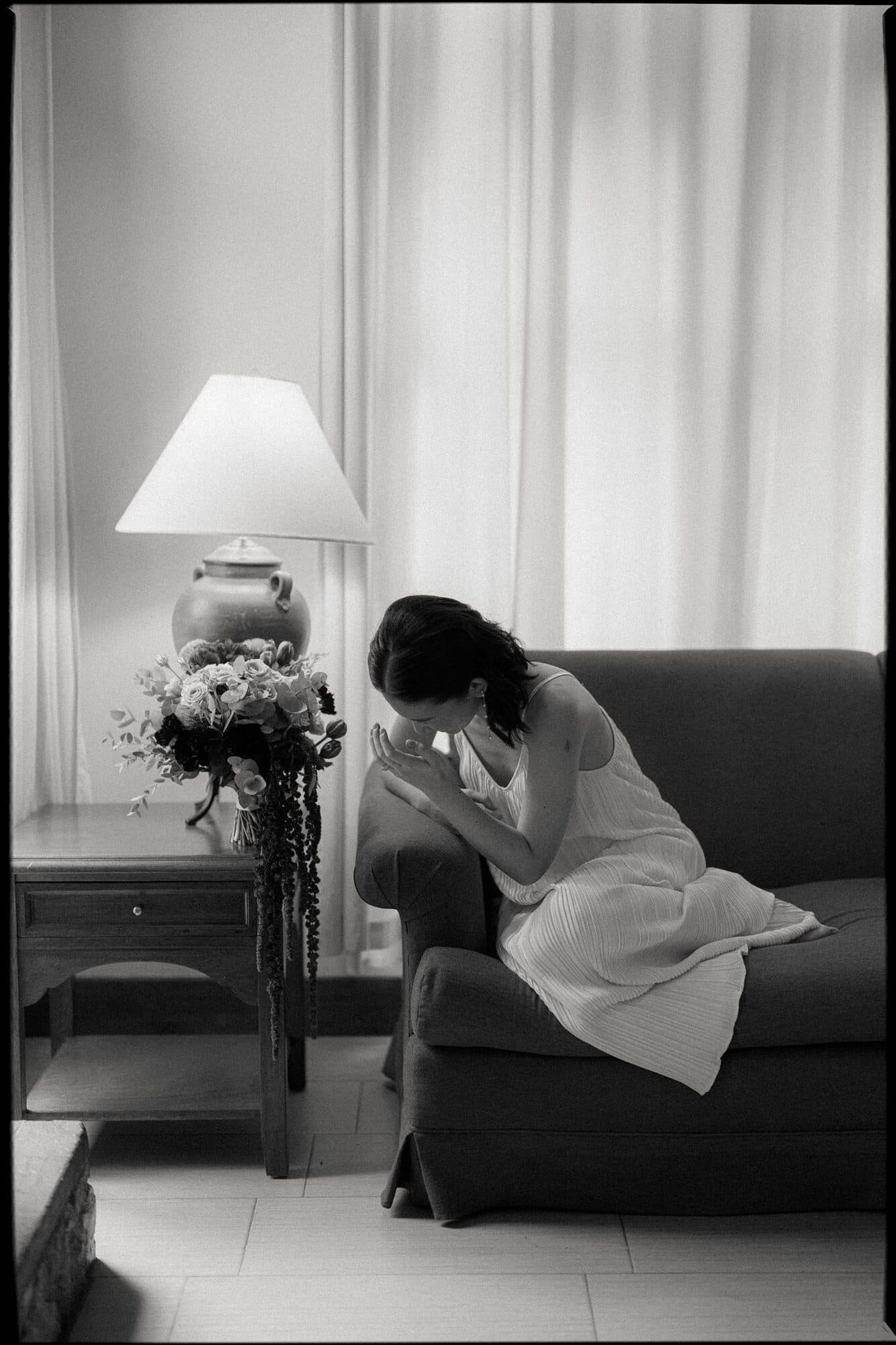 A bride sitting alone on a sofa beside a lamp and her bouquet, head bowed and hands clasped at her face