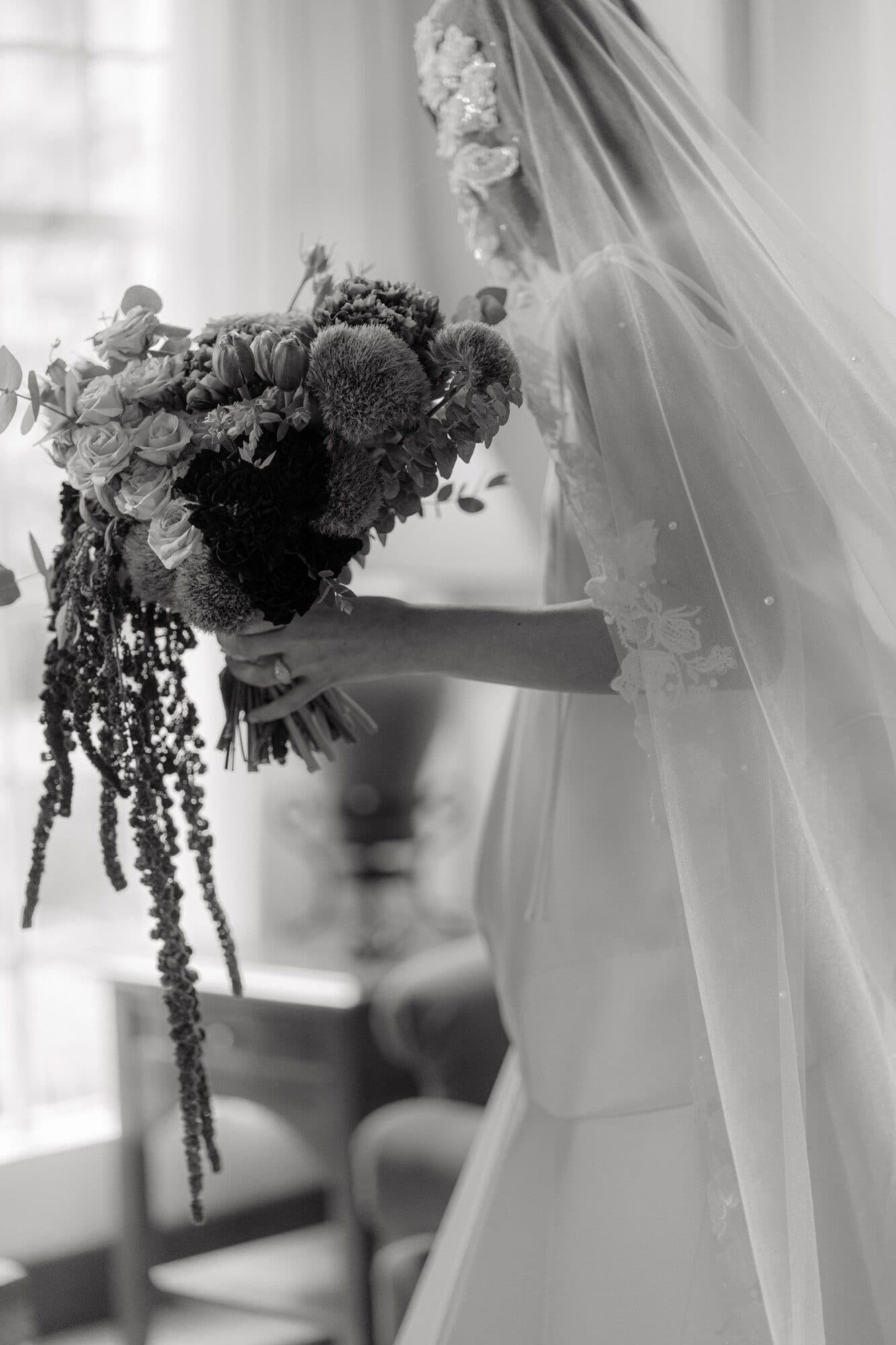 A bride lifting a bouquet of dahlias and trailing amaranthus beside her veil in front of a bright window