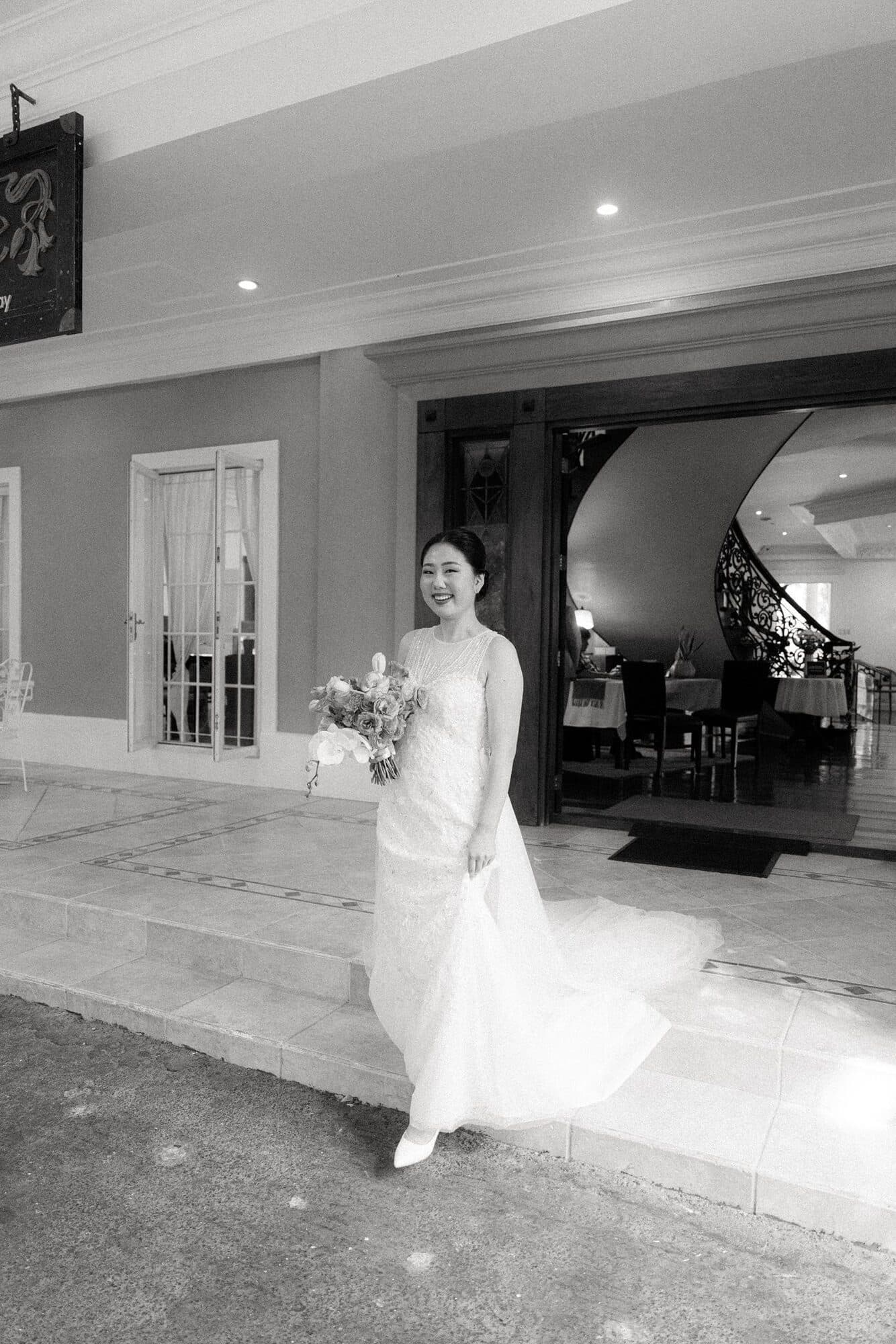 A bride walking down the front steps of a building with her bouquet, the train of her gown sweeping behind her
