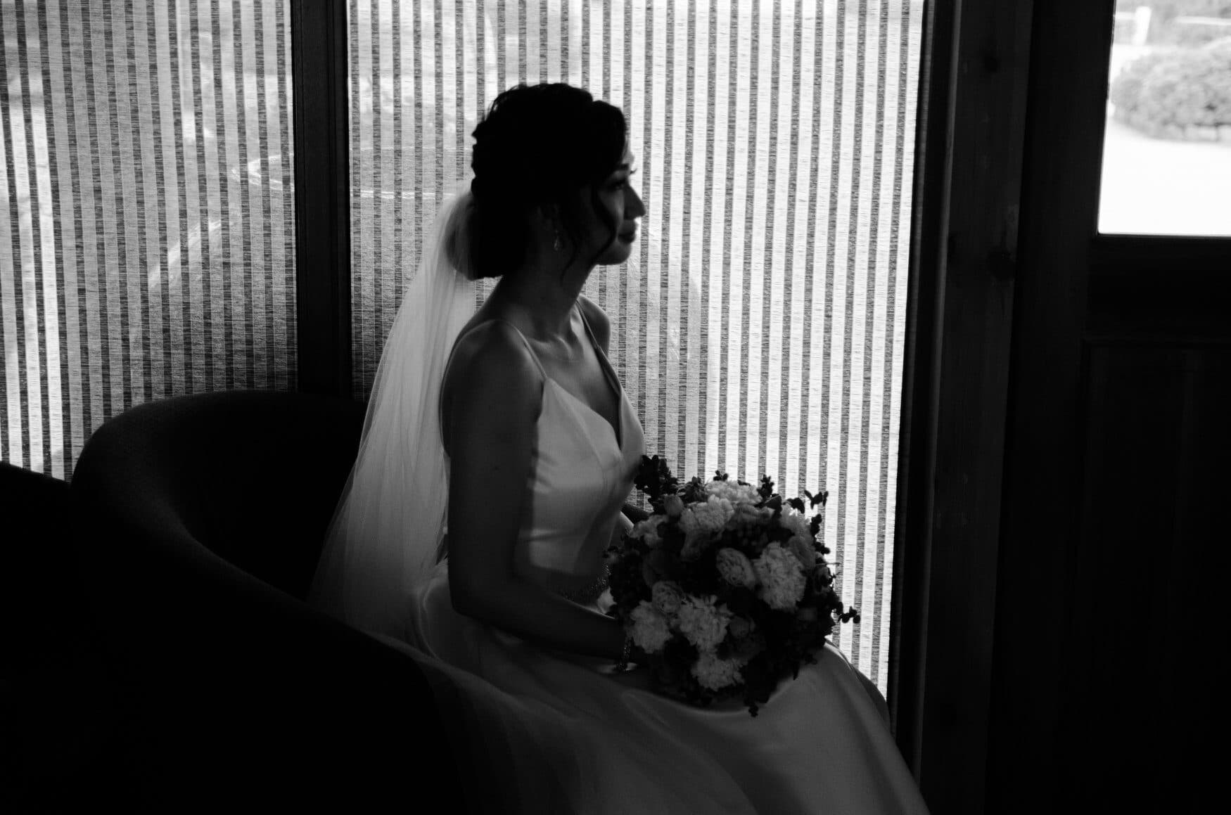 A bride seated in profile by a window, veil over her shoulders and bouquet resting in her lap