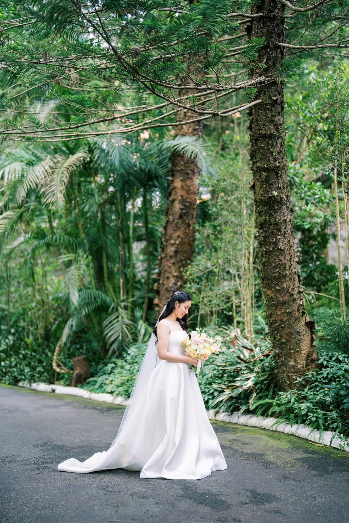 A bride in a full satin gown standing on a driveway framed by pines and tropical ferns, looking down at her bouquet