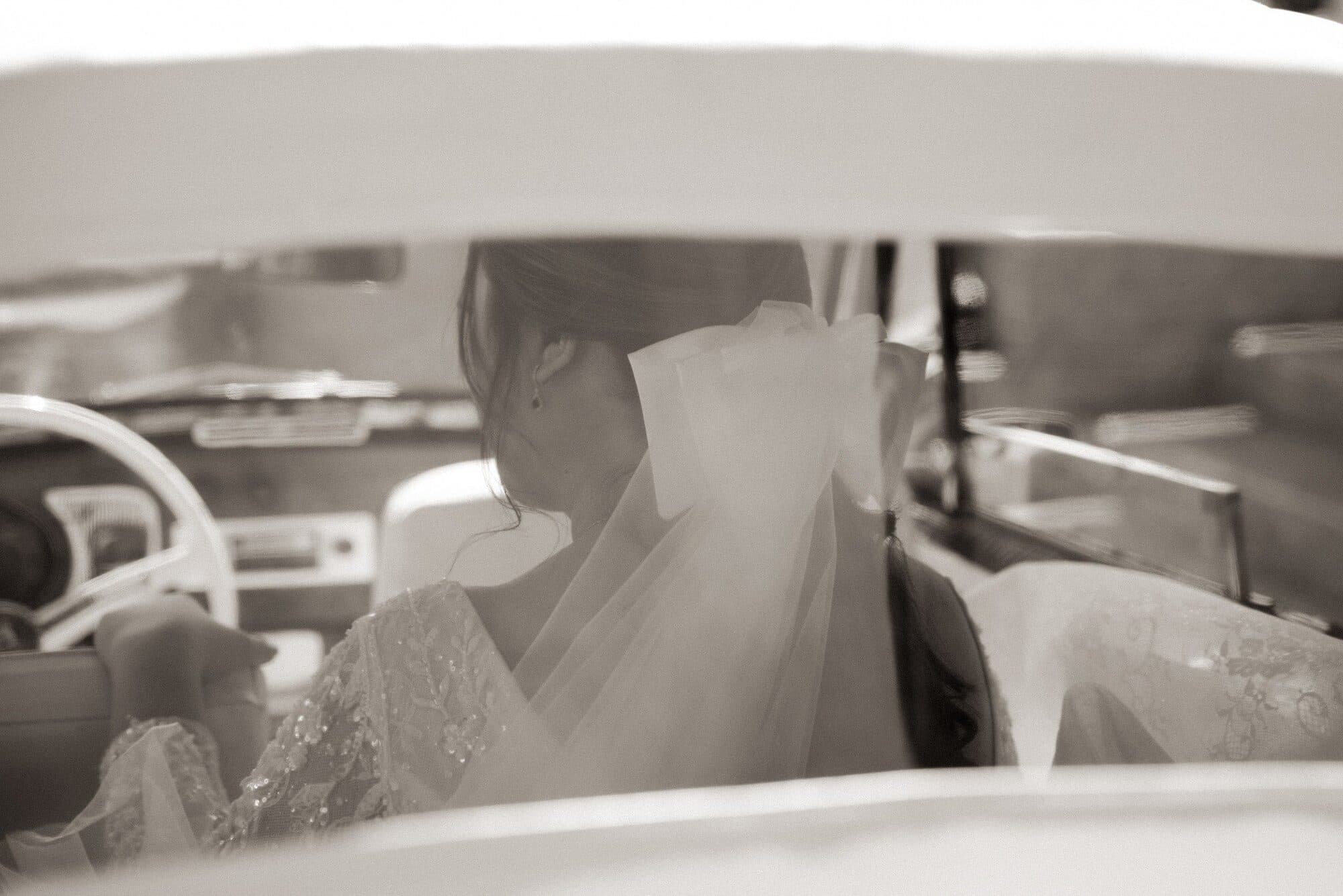 A bride seated inside a car before the ceremony, her veil filling the frame through the windshield