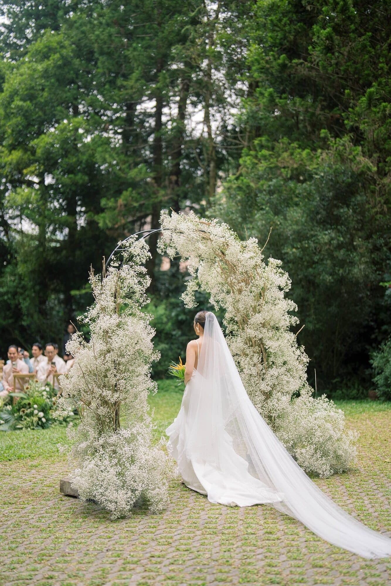 A bride seen from behind at a circular arch of white baby’s breath on a garden lawn, her long veil trailing across the grass as guests look on