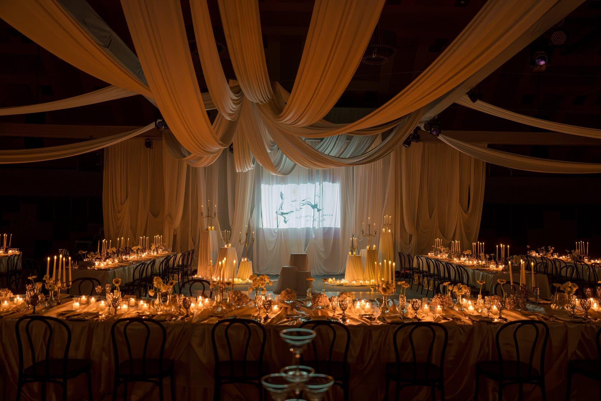 A reception hall draped in soft fabric, with long candlelit tables leading to a lit projection screen