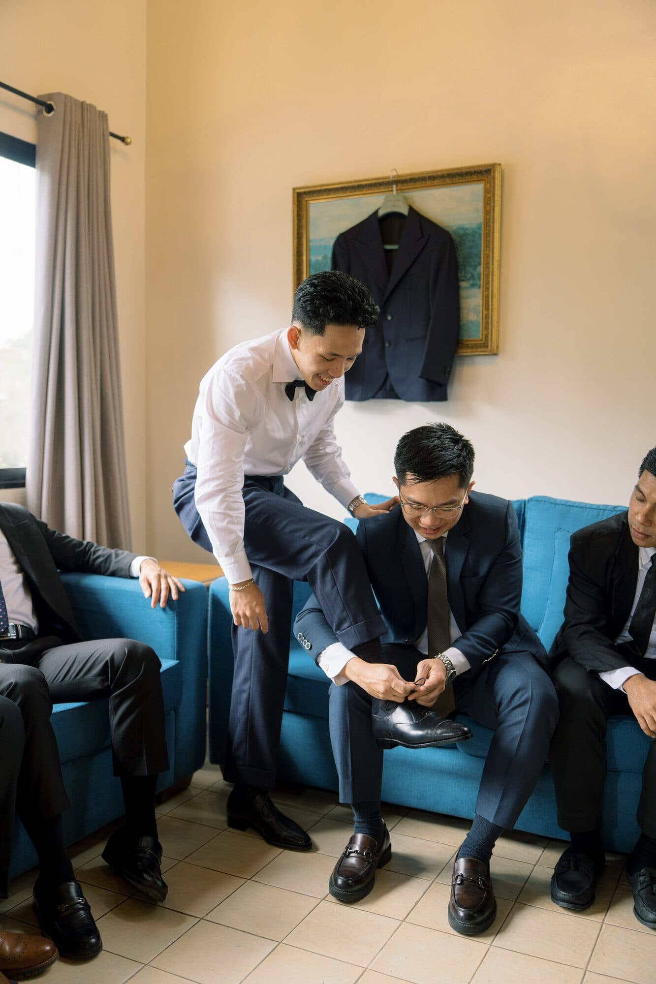 The groom and his groomsmen getting dressed together in a sunlit room, one of them fixing his shoes on a blue sofa