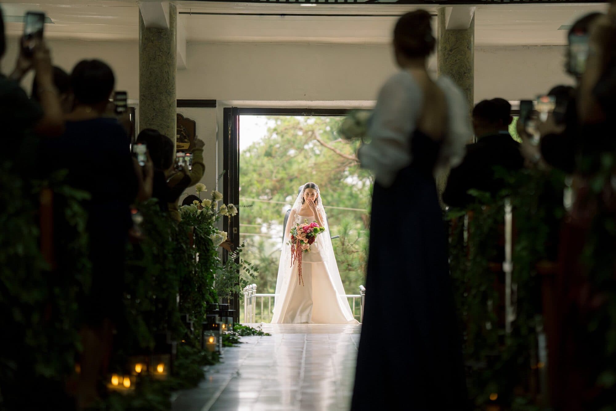 The bride framed in the chapel doorway at the head of the aisle as guests lift their phones to photograph her entrance