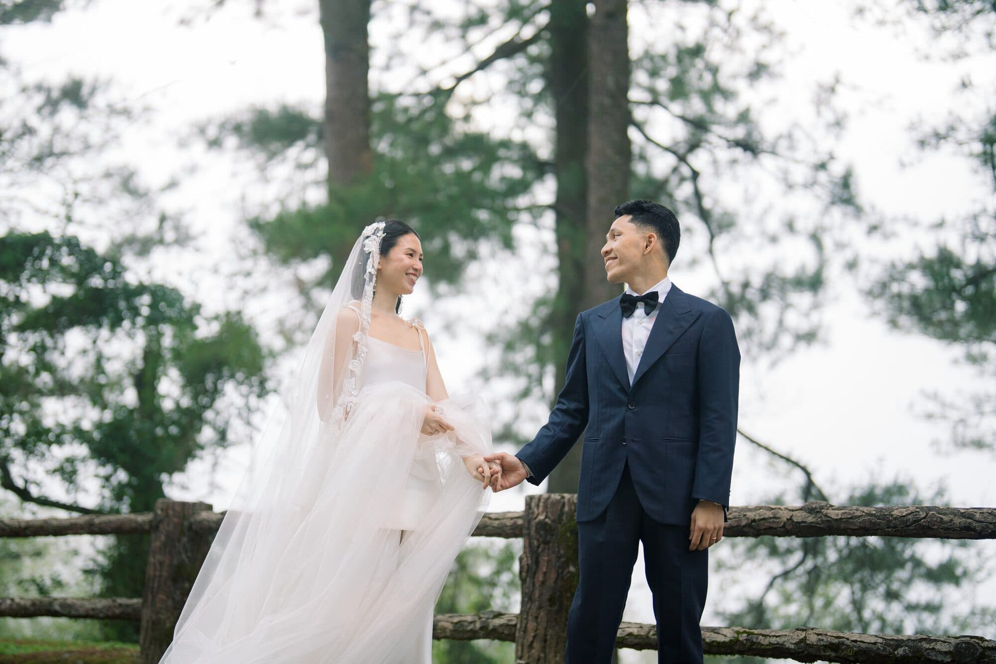 The bride and groom holding hands at a wooden railing with tall pines behind them, smiling at each other