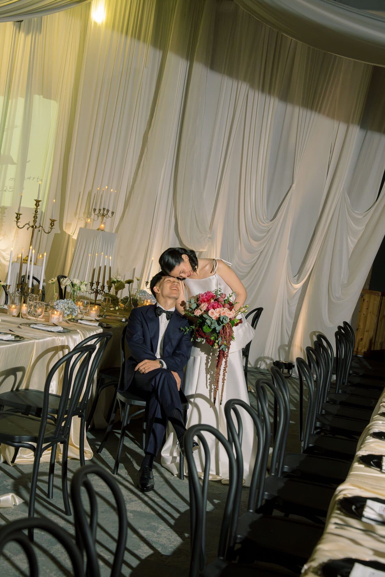 The bride leaning down to kiss the groom at their head table beneath white draped fabric