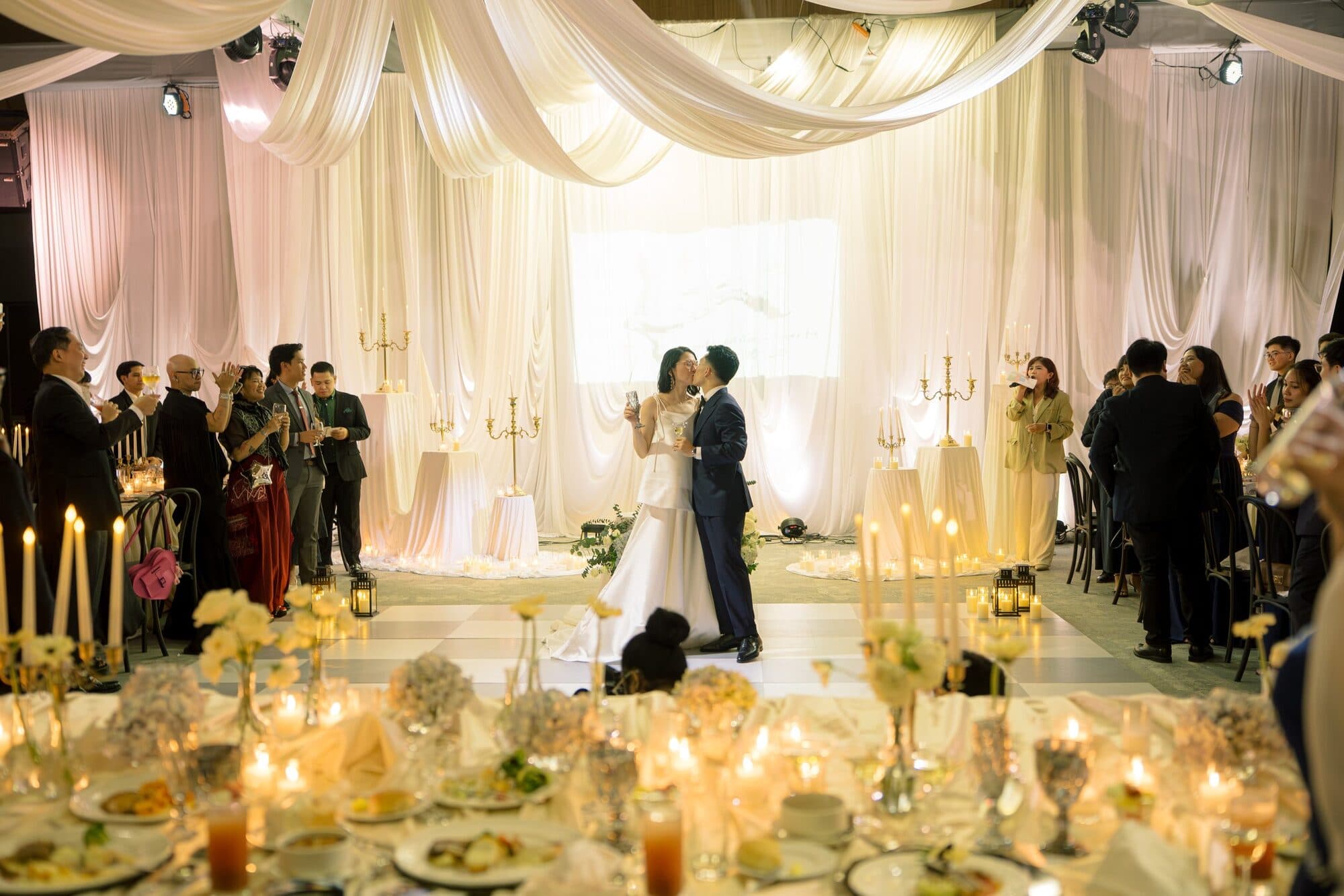 A wide view of the candlelit reception as the couple kiss on the dance floor and guests applaud around them