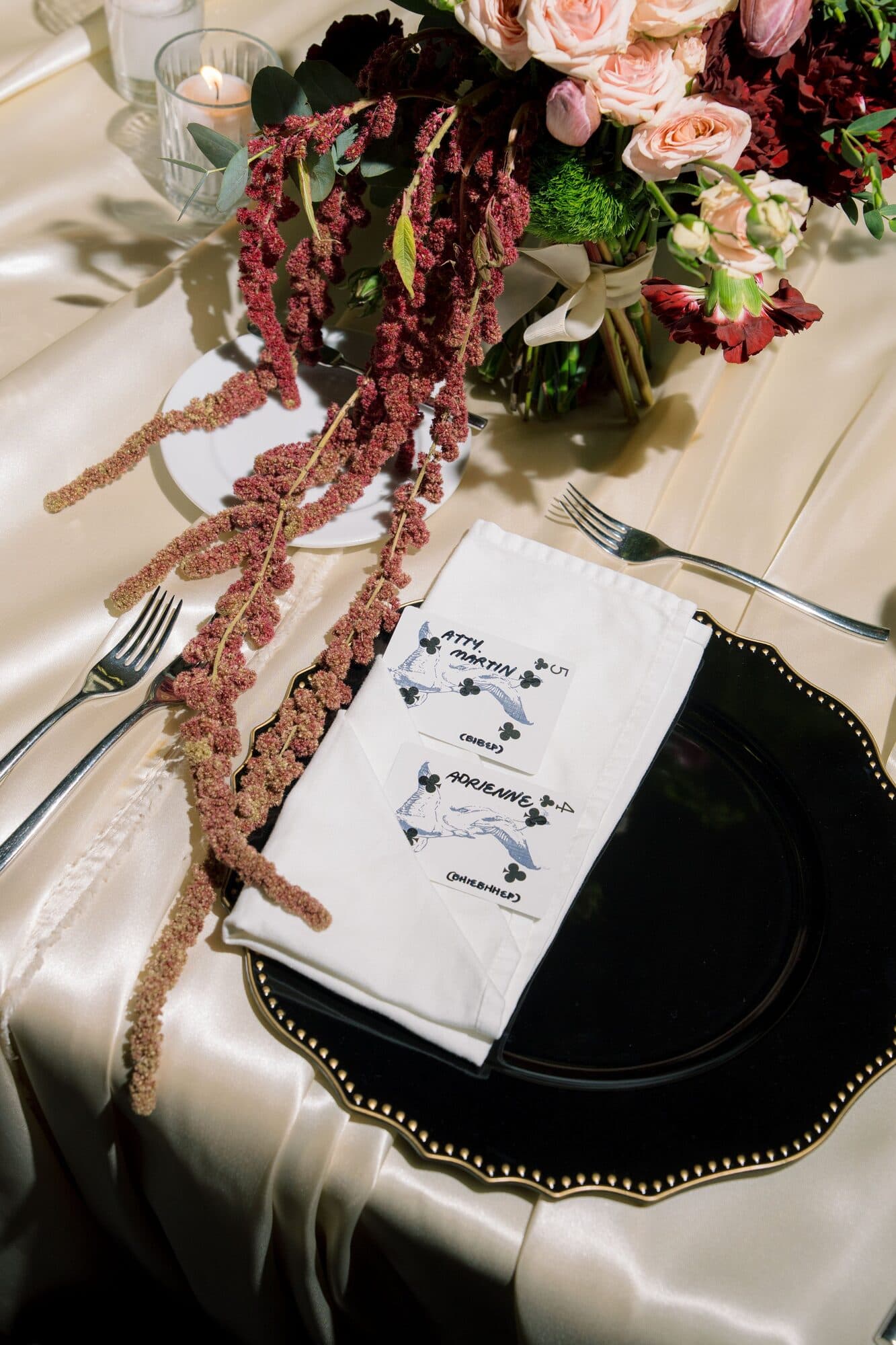A place setting with a printed napkin showing two ducks and the couple’s names, laid on a dark charger beside trailing amaranthus and roses