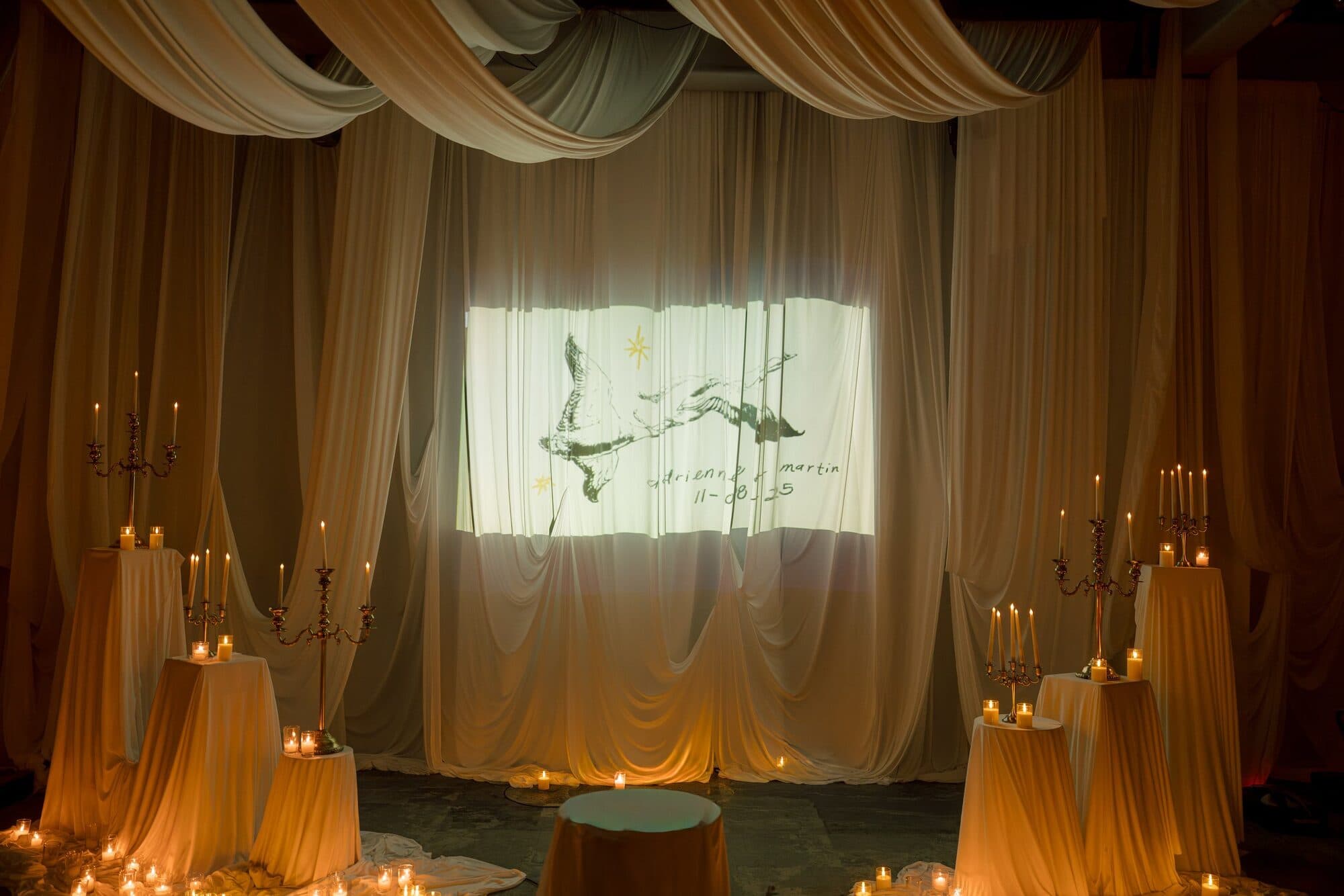The draped reception stage lit by candelabras, with an illustration of two ducks and the couple’s names projected onto the backdrop