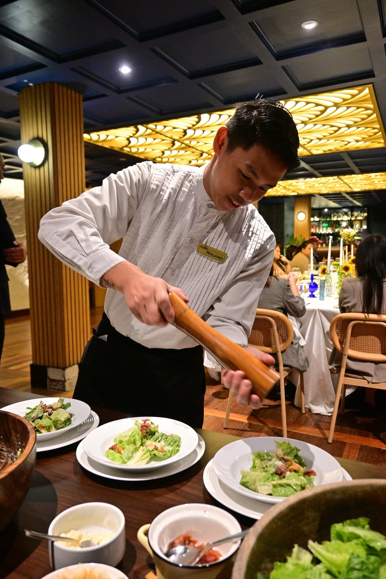 A waiter in a white jacket grinding pepper over plates of Caesar salad prepared tableside