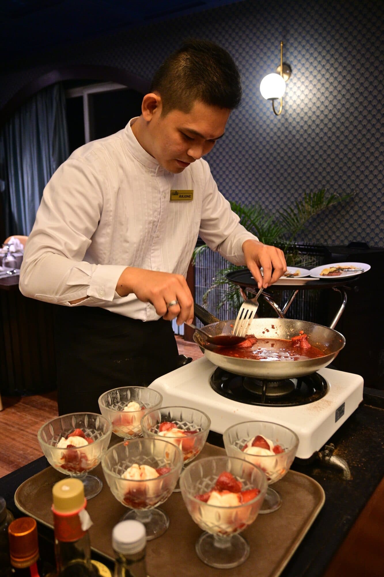 A waiter flambéing the Strawberry Jubilee in a pan over a burner beside a tray of dessert coupes